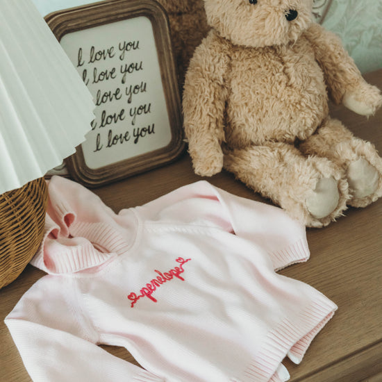 Pink knit sweater for kids personalized with the name "penelope" in dark pink cursive on a dresser