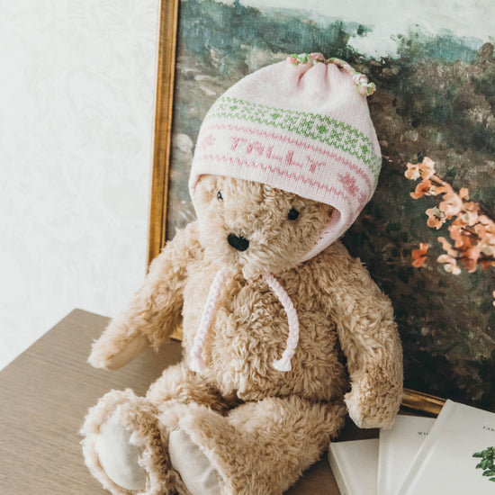 Pink knit hat with pink and green stitching personalized with the name "Tally" worn on a teddy bear